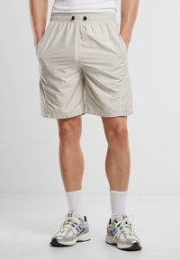 Beige Badehosen aus einem glatten, leichten Stoff. Mit einem elastischen Bund, zwei schwarzen Knöpfen und Seitentaschen.