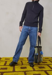 Mörkblå zip-neck tröja, ljusblå raka jeans och en flerfärgad tote-väska med fickor. Texturerad gul och brun matta i bakgrunden.