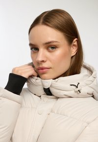 Giubbotto imbottito beige con colletto alto e accenti neri. Presenta una texture trapuntata e bottoni anteriori. Il cordino al colletto offre regolabilità.