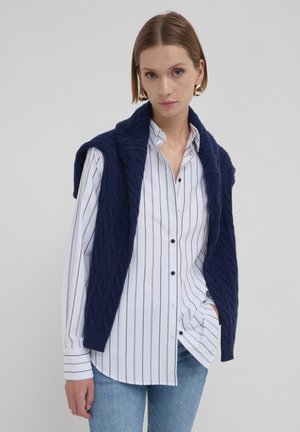 Gilet in maglia a trecce blu navy indossato sopra una camicia bianca a maniche lunghe con righe nere; il modello indossa jeans azzurri chiari e grandi orecchini dorati.