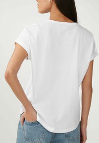 T-shirt en coton blanc avec des manches roulées, un col rond et une coupe décontractée. Détails de coutures visibles au dos et fentes sur les côtés. Porté avec un jean bleu.