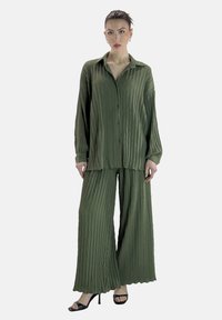 Chemise plissée verte à manches longues et pantalon large assorti, tous deux ornés d'un motif zigzag texturé, associés à des escarpins noirs.