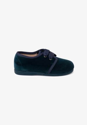 Scarpe in velluto verde scuro con punta arrotondata, fasce elastiche blu navy e suola in gomma. Texture liscia con dettagli decorativi minimi.