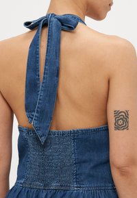 Jeans halterneck kjole med knyttet bak, smocket liv og slaktskjørt. Tøyet er i mellomblått med en glatt tekstur.