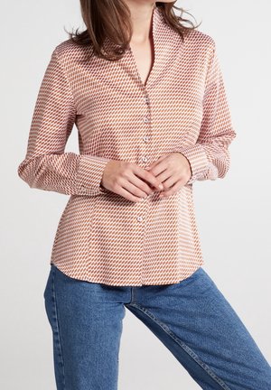 Donna che indossa una camicia marrone e bianca a maniche lunghe con motivo geometrico e jeans blu, mani intrecciate in vita.