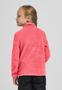 Pullover in pile rosa con colletto alto, superficie texturizzata e maniche lunghe. Il modello indossa pantaloni neri, girato di spalle rispetto alla fotocamera.