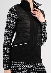 Giacca sportiva nera con torso imbottito, maniche a fantasia con disegni geometrici bianchi e tasche con zip. Design con colletto alto.