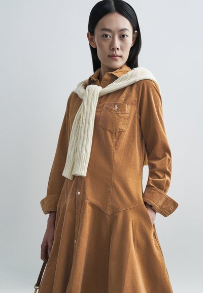 Langärmeliges, knöpfbares Kleid aus rotem Cordstoff mit einer Brusttasche und einem cremefarbenen Zopfmuster-Pullover, der über den Schultern drapiert ist.