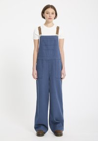 Blauwe denim salopettes met wijde pijpen, voorzien van verstelbare bruine banden, een voorzak en twee zijzakken. Gemaakt in combinatie met een wit shirt.