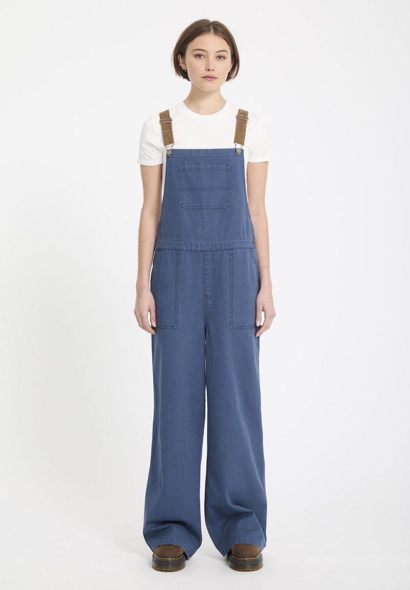Blauwe denim salopettes met wijde pijpen, voorzien van verstelbare bruine banden, een voorzak en twee zijzakken. Gemaakt in combinatie met een wit shirt.