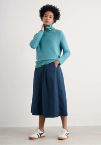 Un pull côtelé turquoise superposé à un col roulé rayé vert et blanc, associé à une jupe culotte marine et des baskets blanches avec des accents verts.
