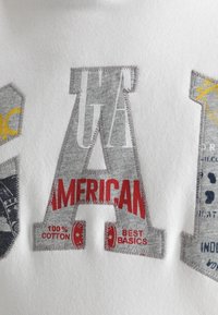 Šedá látková nášivka ve tvaru písmene "A" s červeným textem "AMERICAN," "100% COTTON," a "BEST BASICS" vyšitým na bílém textilním pozadí.