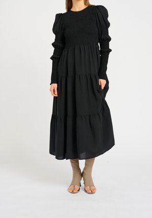 Kobieta ubrana w długą czarną sukienkę z falbanami, z gorsetem marszczonym i bufkami na długich rękawach, do tego beżowe botki na obcasie z odkrytymi palcami.