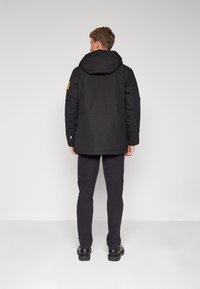 Veste isolante noire avec capuche, présentant un extérieur lisse, des poignets réglables et un patch logo sur le haut du bras. Associée à un pantalon foncé.