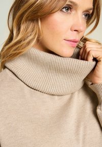 Beżowy, dziergany sweter z golfem i prążkowanym kołnierzem. Miękka faktura, dopasowany krój i gładka tkanina, częściowo trzymana ręką osoby noszącej.