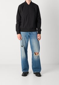 Schwarze Bomberjacke mit Reißverschluss vorne, gerippten Bündchen und Saum; kombiniert mit hellblauen, weit geschnittenen Jeans mit zerrissenen Knien und schwarzen Schuhen.