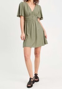Abito verde oliva con scollatura a V, maniche corte a sbuffo e vita arricciata. Presenta una gonna fluida. Sandali neri con cinturino completano l'outfit.