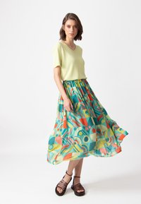 Limoengroen V-hals top gecombineerd met een kleurrijke, gestructureerde maxi rok met geometrische ontwerpen in oranje, teal en geel. Bruine sandalen met banden.