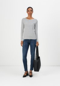 Top à rayures à manches longues en noir et blanc, associé à un jean ajusté bleu. Le mannequin tient un sac fourre-tout noir et porte des chaussures à talons noirs.