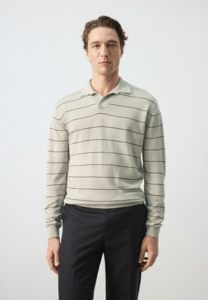 Un homme se tient face à l'avant, portant un polo à manches longues gris clair avec de fines rayures horizontales noires et un pantalon foncé, sur un fond uni.