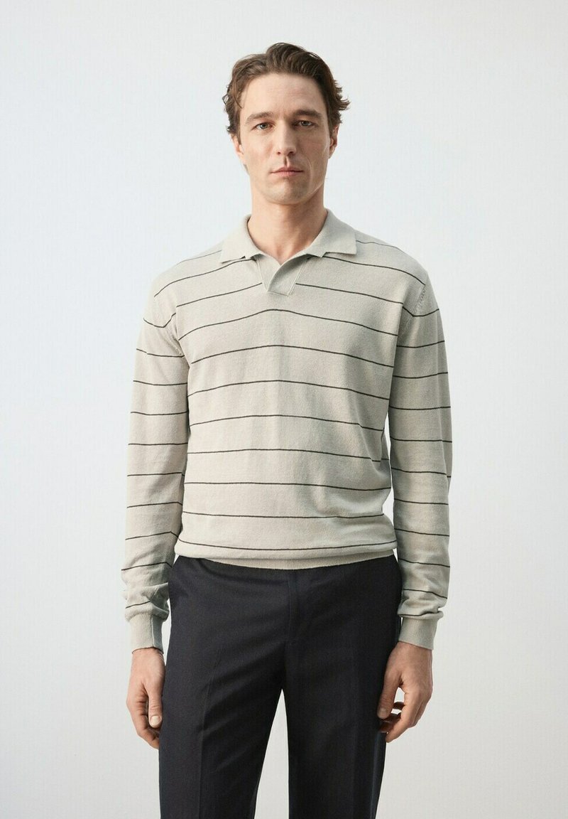 Un homme se tient face à l'avant, portant un polo à manches longues gris clair avec de fines rayures horizontales noires et un pantalon foncé, sur un fond uni.