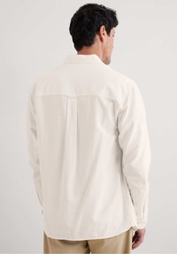 Ljusbeige långärmad skjorta med klassisk krage, knäppta manschetter och rundad nederkant, med en centralt bakveck och mjuk textur.