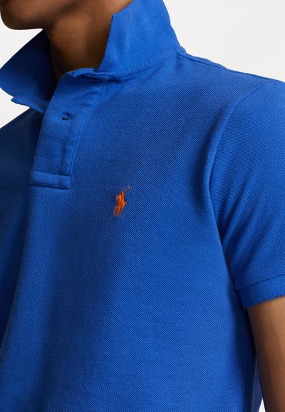 Camisa polo azul hecha de una tela texturizada, con un cuello corto, tapeta de tres botones y un logo bordado naranja en el pecho.