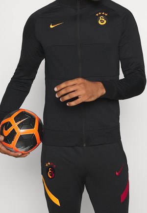 Personne en tenue de sport noire avec le logo Galatasaray tient un ballon de football Nike noir et orange sur un fond uni.