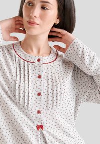 Camiseta de pijama de color gris claro con un patrón que presenta pequeños diseños florales, un cuello redondo con ribete rojo, cierres de botón y un detalle de lazo en el dobladillo.