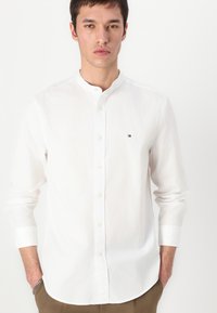 Camicia bianca con colletto alla mandarina, caratterizzata da bottoni gialli e un piccolo logo ricamato sul petto. Il tessuto è liscio e leggero.