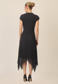 Vestido negro con pliegues, mangas cortas tipo capucha, dobladillo asimétrico y un acabado liso en el cuerpo. Lleva consigo unos zapatos de tacón alto negros que exhiben una tela texturizada.