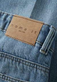 Denimstoff i lyst blått med en lærmerke sydd på, som viser "name it" og "EST. 1986" i brun tekst. Synlige sømmer.