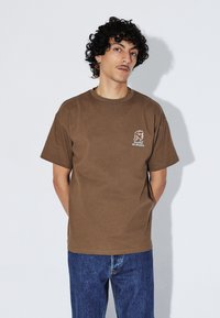 T-shirt en coton marron à manches courtes, avec un graphisme blanc représentant un profil et le texte "Rêves d'Utopie" sur la partie gauche de la poitrine.