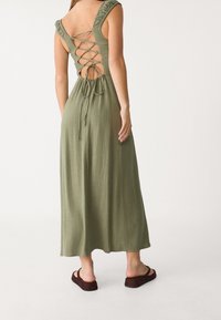 Vestido maxi verde oliva con tirantes de hombro con volantes, espalda descubierta con detalle de cordones y cintura ajustable. Llevado con zapatos marrones de tipo slip-on.