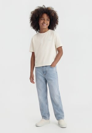 Kind met krullend haar lacht, draagt een crèmekleurig T-shirt met korte mouwen, lichtblauwe spijkerbroek en witte slip-on schoenen, staat met één hand in de zak.