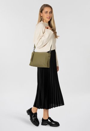 Borsa a tracolla in pelle verde oliva con finitura texture e nappine decorative. Il modello indossa un maglione crema e una gonna plissettata nera.