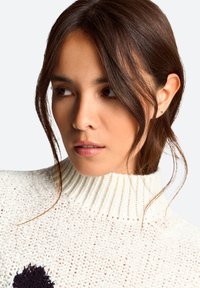 White knit sweater with a high ribbed collar and black accent pattern. Smooth texture and loose fit, featuring a chunky knit design.
