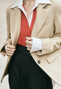 Beige uld blazer med en enkelt knap, lagdelt over en rød striktop med en hvid krave, parret med højtaljede sorte bukser.