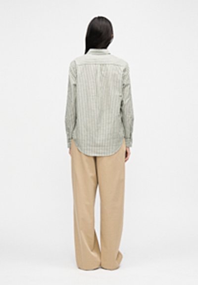 Pruhovaná košeľa na gombíky v svetlozelených a bielych farbách, s dlhými rukávmi a lemom, ktorý sa nahor zakrivuje, v kombinácii so svetlobéžovými širokými nohavicami.