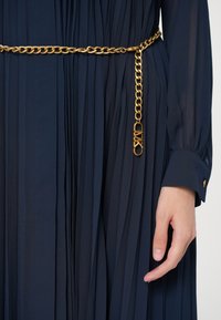 Ναυτικό μπλε πλισέ φόρεμα με μακριά μανίκια με προσαρμοσμένα μανίκια, συνδυασμένο με μια χρυσή αλυσίδα μέσης που διαθέτει μενταγιόν λογότυπο, δίπλα σε μια χαλαρή χερι.