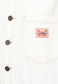 Balts džinsu krekls ar kabatu. Iespējas ietver oranžas šuves un taisnstūrveida "AMV DENIM" etiķeti ar palmu un saulrieta dizainu. Melni pogas.