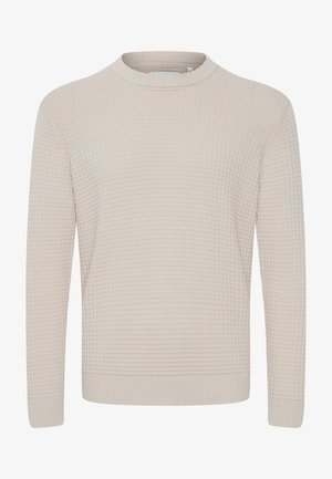 Uomo con un maglione beige a trama, jeans scuri e sneaker nere in piedi contro uno sfondo chiaro e semplice.