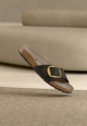 Μαύρο σανδάλι slide με μεγάλη χρυσή αγκράφα, φελλό πάτο και υφασμένο πάτο τοποθετημένο σε χαλί μπροστά από μπεζ καναπέ.