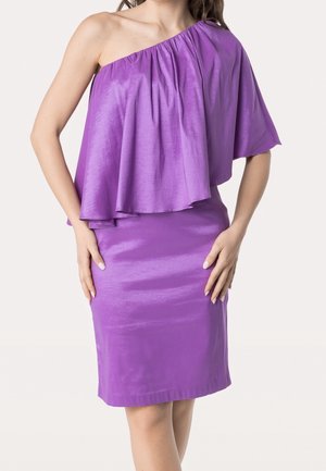 Žena v lesklých fialových šatech s jedním ramínkem, s plápolajícím róbovým překrytím a přiléhavou sukní po kolena, ruce opřené o boky.