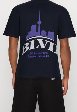 Camiseta de algodón azul marino con un gran gráfico de un horizonte de ciudad en púrpura y el texto “BLVD” en blanco sobre el horizonte.