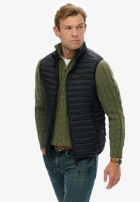 Chaleco de plumas negro sobre un suéter de punto trenzado verde. Presenta un alto cuello, cierre de cremallera y un pequeño logo. Combinado con jeans azules y un reloj.