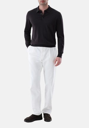 Mann trägt schwarzen langärmeligen Poloshirt, weiße Hose und dunkelbraune Schuhe, steht mit einer Hand in der Hosentasche vor weißem Hintergrund.