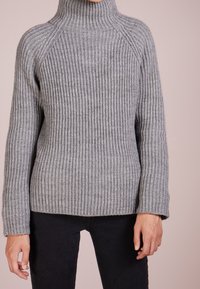 Eine Person, die einen dicken, gerippten grauen Rollkragenpullover und schwarze Hosen trägt, steht vor einem hellrosa Hintergrund.
