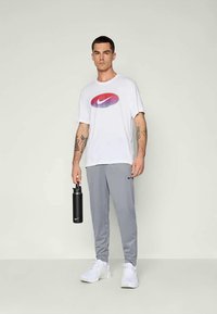 Meeste valge Nike t-särk lilla logoga, hallid sportpüksid ja valged spordijalatsid. Hoides musta veepudelit, seistes heleda taustaga.