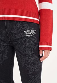Roter Pullover mit weißen Streifen an den Ärmelbündchen, kombiniert mit schwarzen Jeans mit kunstvollen tonalen Designs und einem weißen Logo.